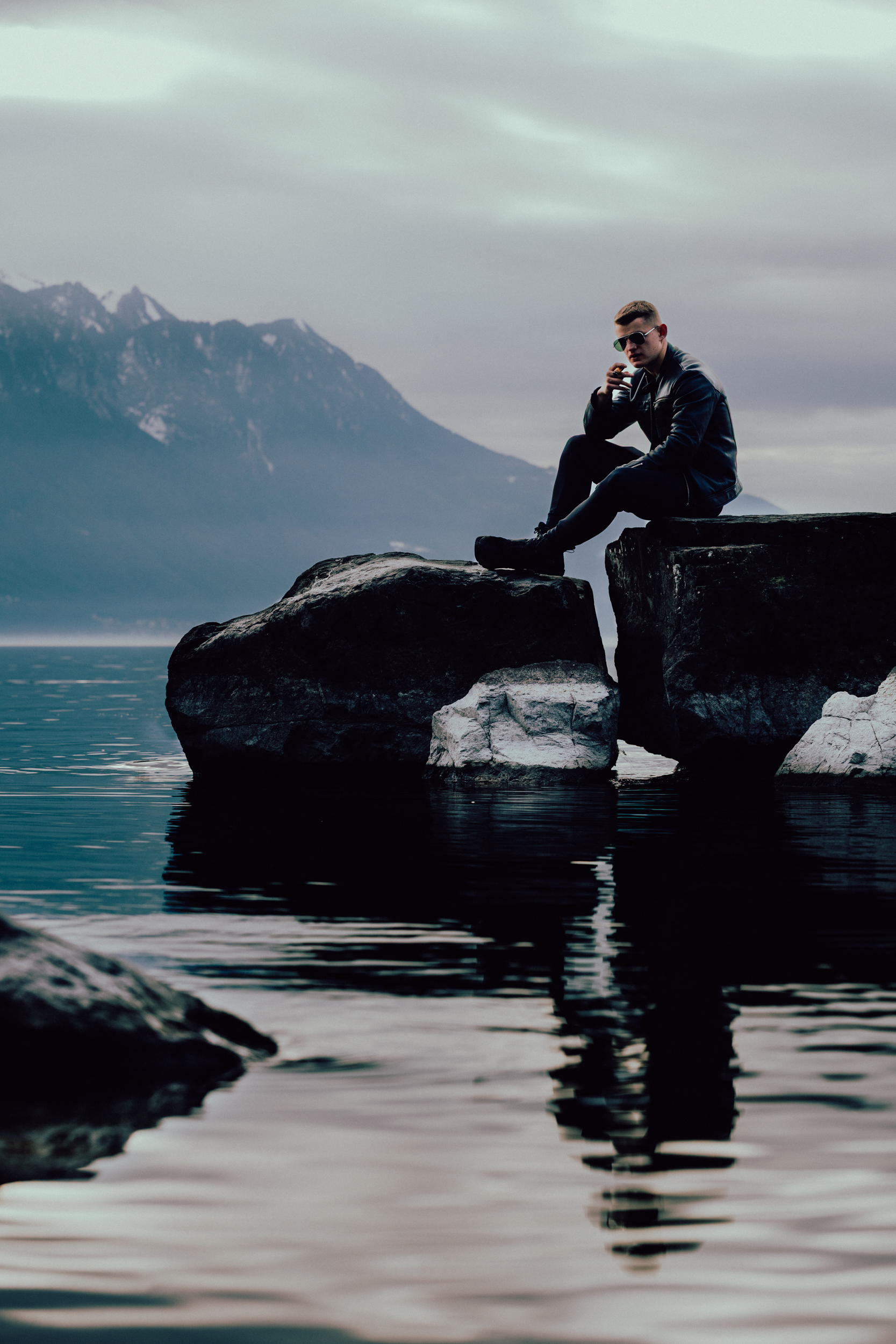 R 5 man in leather jacket smoking cigar on rocks at Lake Geneva